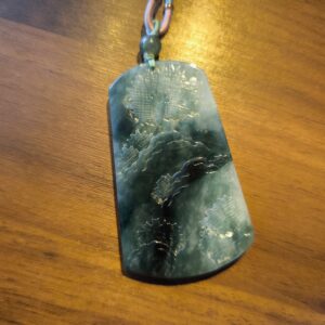 This is a beautifully crafted jade pendant featuring deep green and translucent hues, creating a striking natural gradient across its surface. The piece is meticulously carved with intricate designs, likely depicting traditional Chinese motifs such as pine trees, mountains, and possibly cranes or phoenixes — symbols of longevity, strength, and peace. The pendant is polished to a glossy finish and suspended from a decorative cord, complemented by a round jade bead at the top. Its elegant form and cultural symbolism make it both a meaningful keepsake and a refined accessory.